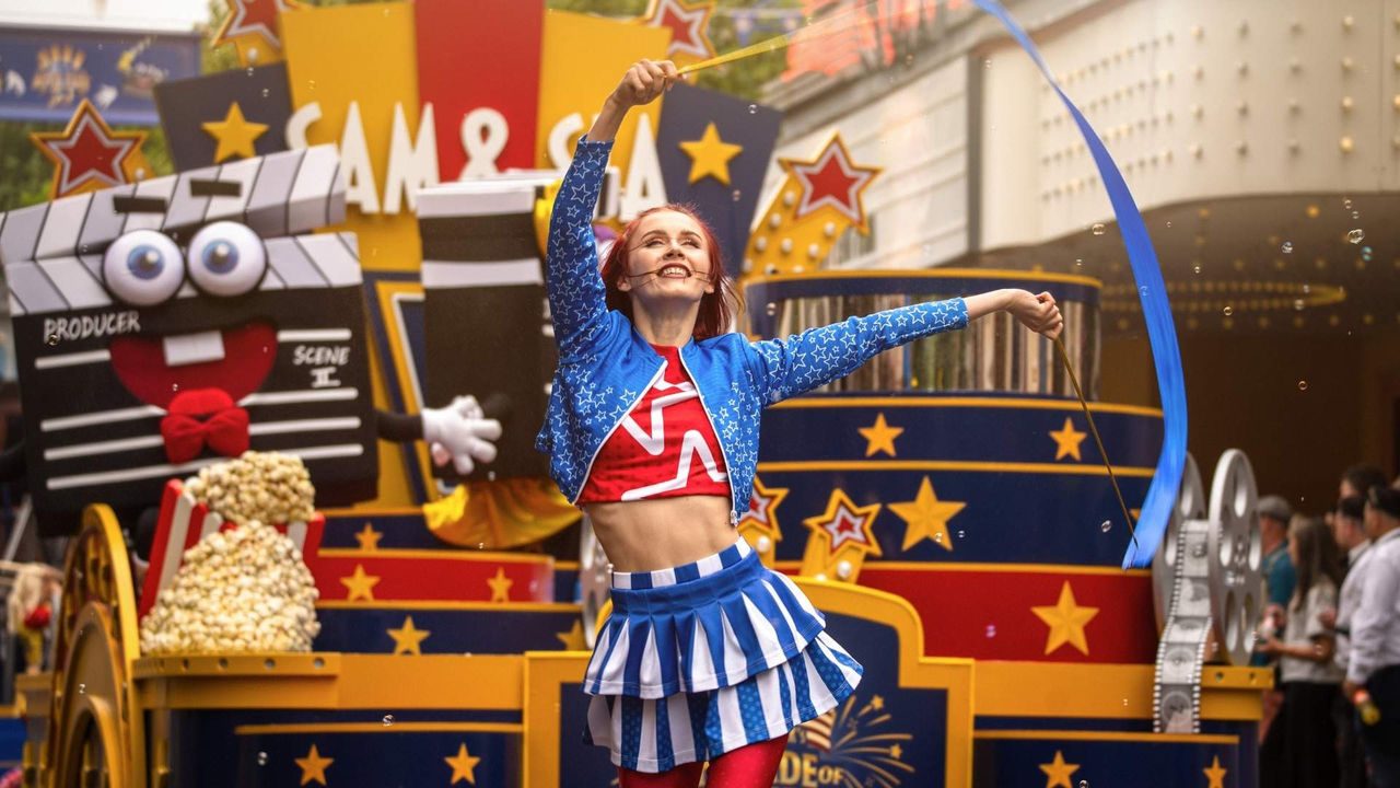 Person in einem blau-rot-weißen Showkostüm schwingt bei einer Parade ein langes blaues Band vor einem bunt dekorierten Wagen mit Film-Thema. Im Hintergrund sind Filmrollen, Sterne, Popcorn-Dekorationen und eine Comic-Klappe mit großen Augen zu sehen, während Zuschauer am Straßenrand stehen.