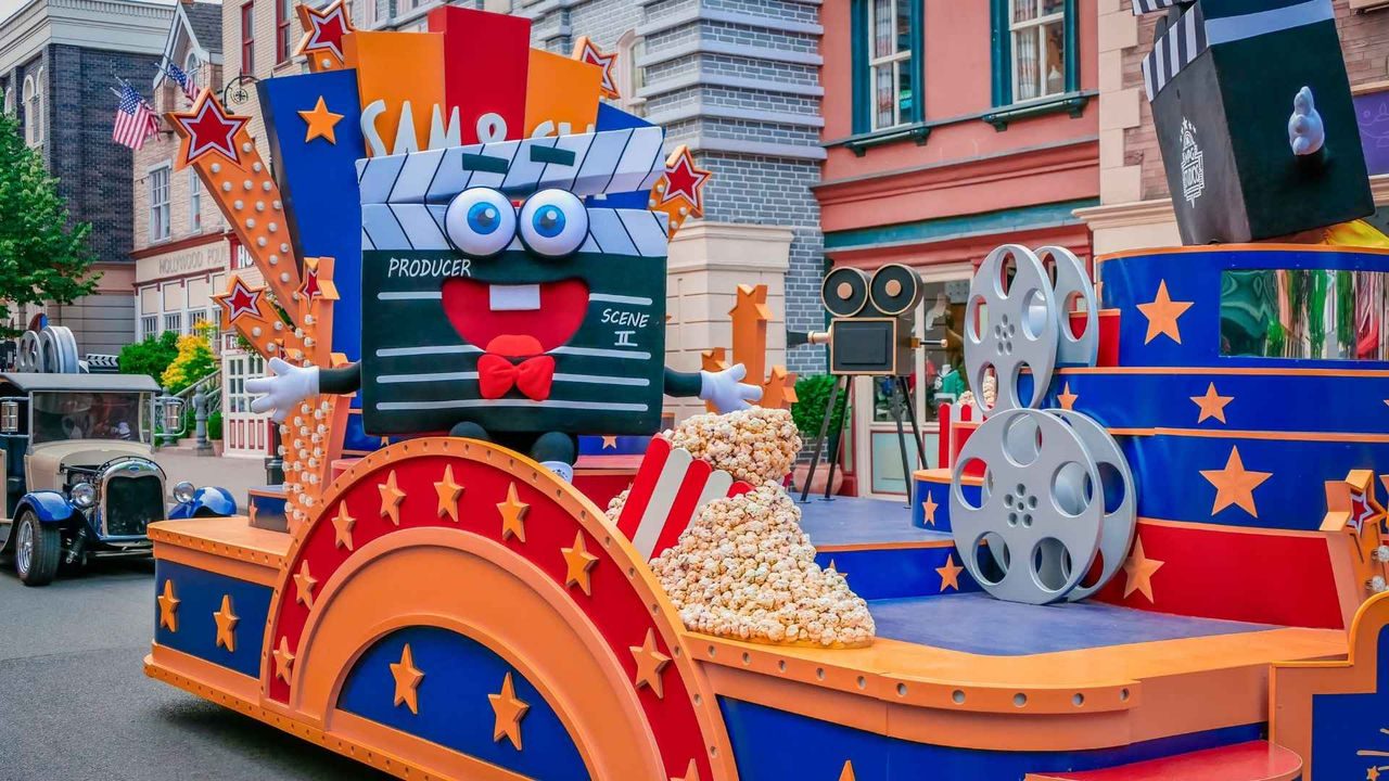 Bunter Themenpark-Paradenwagen im Filmstil mit großer, lächelnder Filmklappe („Producer“, „Scene II“), Filmrollen, Popcorn-Dekoration und Sternmotiven in einer Straßenszenerie mit Gebäuden im Hintergrund.
