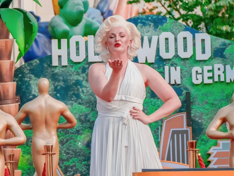 Person in a white pleated Marilyn Monroe–style dress stands in front of a “Hollywood” backdrop and blows a kiss, surrounded by golden statues, palm elements, and a red carpet.