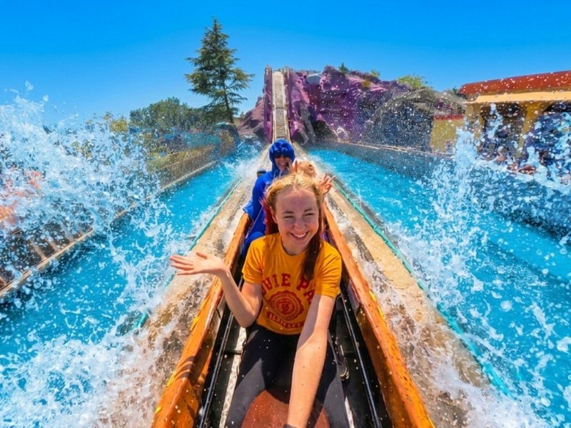 A person is sitting in the front seat of a water ride and raises a hand as a large wave splashes up on both sides. Another person is sitting behind them in the boat. In the background, you can see the ride’s steep descent, a colorful rocky landscape, and a sunny, blue sky.