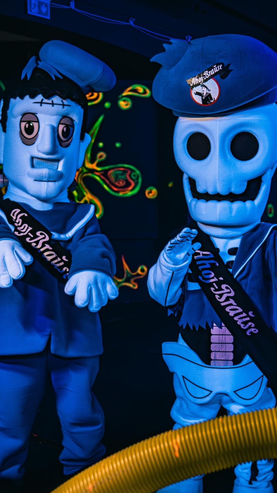 Frankie en Skulli uit het Ahoj-Brause Horror Lab staan in een laboratoriumomgeving die wordt verlicht door blacklight. De twee figuren dragen blauwe matrozenpetten met het Ahoj-Brause-logo en kijken recht in de camera. Op de donkere muren op de achtergrond lichten kleurrijke fluorescerende patronen op.