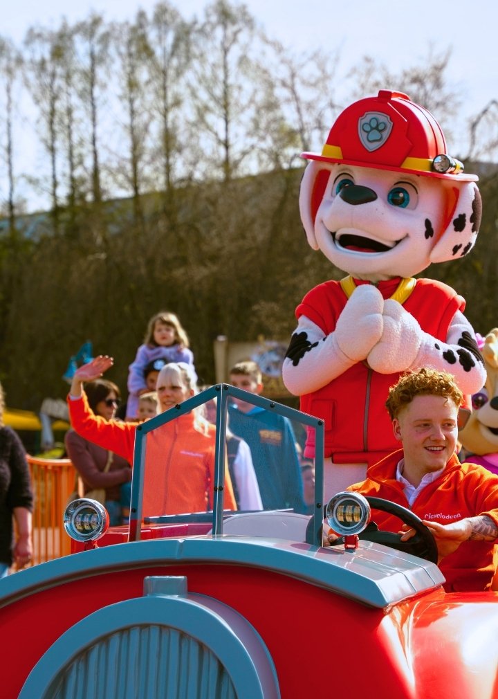 Eine farbige Grafik zeigt eine animierte Figur aus dem PAW‑Patrol‑Universum in einer Sprungbewegung im Vordergrund. Die Figur hat ein türkisfarbenes Outfit und eine helle Mütze. Im Hintergrund ist eine hügelige Landschaft mit Wegen und Bäumen zu sehen sowie ein hoher Turm mit einer runden Plattform oben. Der Himmel ist blau mit mehreren strahlenförmigen Lichteffekten.