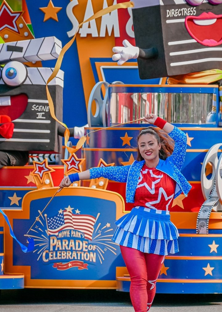 Bunte Parade in einem Freizeitpark: Ein festlich dekorierter Wagen mit Filmklappen-Figuren, Filmrollen, Popcorn und Sternenmotiven fährt über eine Parkstraße. Im Vordergrund tanzt eine Person in einem rot-blau-weißen Kostüm mit langen Bändern. Auf dem Wagen ist das Logo „Movie Park’s Parade of Celebration“ zu sehen. Weitere Paradefahrzeuge folgen im Hintergrund, umgeben von bunten Gebäudefassaden, Bäumen und sonnigem Wetter.