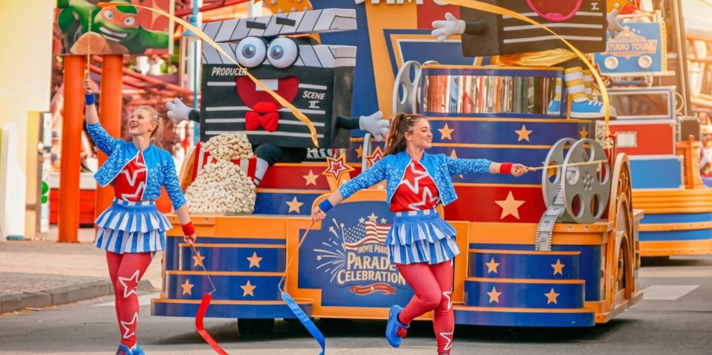 Bunte Parade in einem Freizeitpark: Ein festlich dekorierter Wagen mit Filmklappen-Figuren, Filmrollen, Popcorn und Sternenmotiven fährt über eine Parkstraße. Im Vordergrund tanzt eine Person in einem rot-blau-weißen Kostüm mit langen Bändern. Auf dem Wagen ist das Logo „Movie Park’s Parade of Celebration“ zu sehen. Weitere Paradefahrzeuge folgen im Hintergrund, umgeben von bunten Gebäudefassaden, Bäumen und sonnigem Wetter.