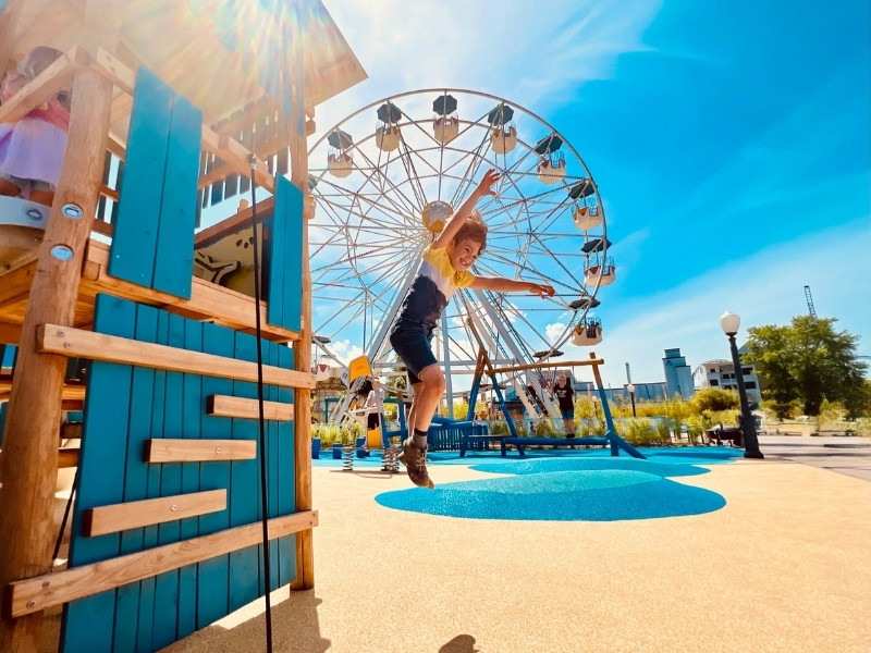 Ein Kind springt von einem Klettergerüst auf einem Spielplatz. Im Hintergrund sieht man ein Riesenrad.