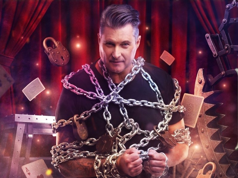 Promotional graphic for the show “100 Years Houdini”: A person stands on a stage set with red curtains and is wrapped multiple times in metal chains and padlocks. Playing cards and props from an escape and magic show float around the person, including locks, a large saw, and a suspended restraint device. On the right side of the image, the title “CHRISTIAN FARLA – 100 YEARS HOUDINI” is displayed in large, metallic-looking letters. The scene is bathed in warm, dramatic lighting and conveys the atmosphere of a classic illusion show.