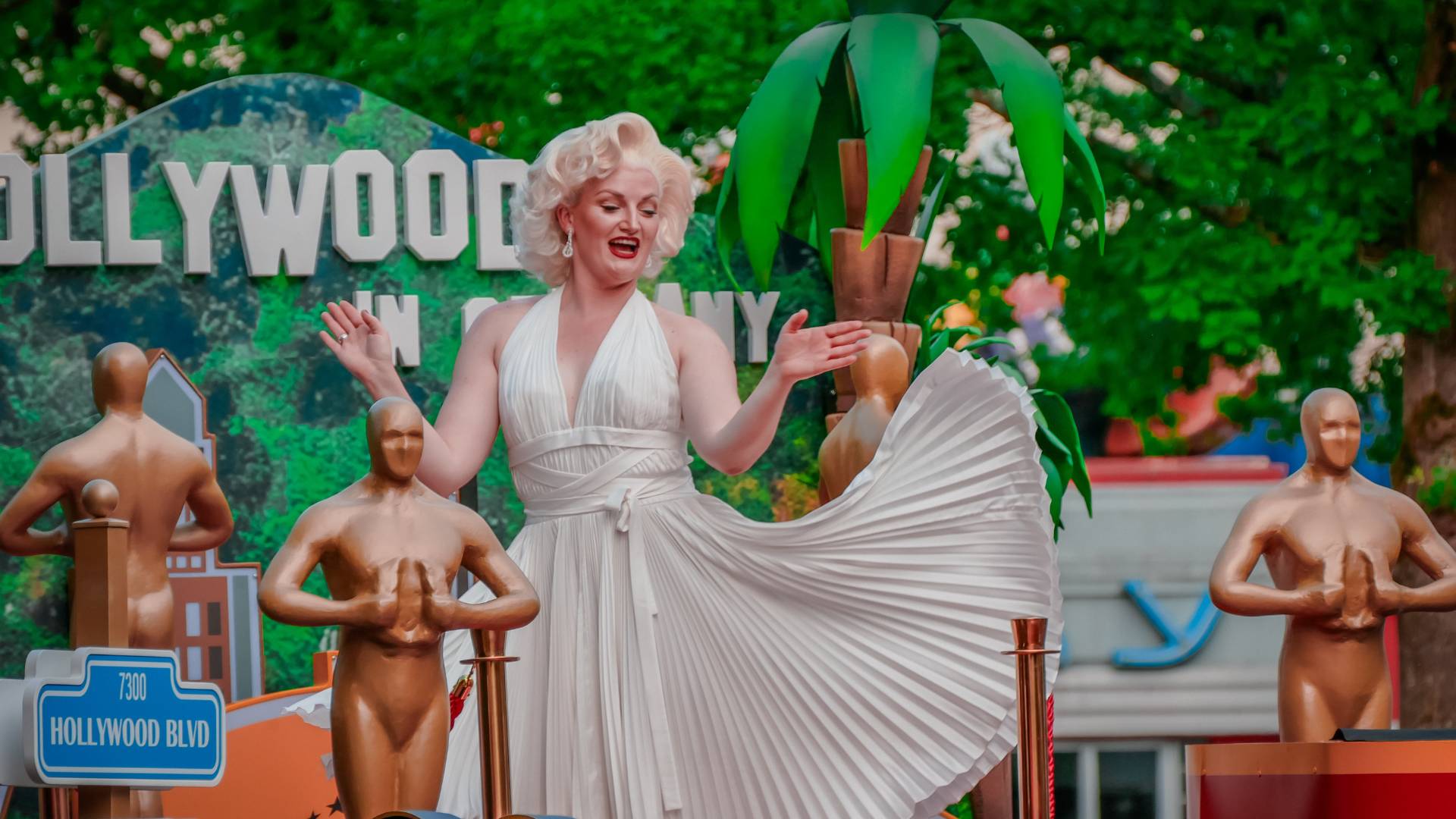 A group of several individuals is moving along a street within a theme park area. In the foreground, one individual wearing a light-colored, fringed costume is moving toward the camera. Behind this individual, others follow in formal or performance-style clothing, including outfits with suspenders and bow ties. In the background, themed buildings, a yellow vehicle, and a large decorative film reel structure are visible. The scene takes place outdoors in daylight.