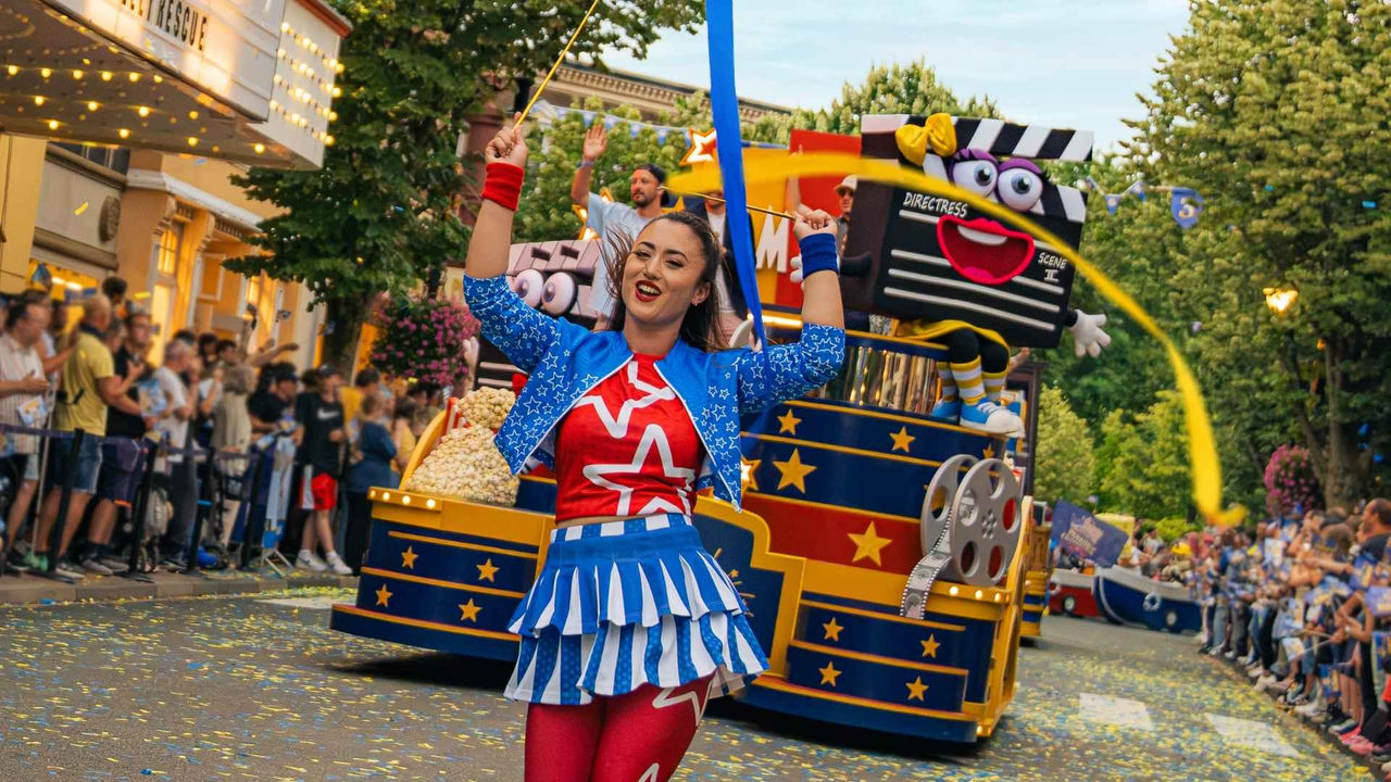 Person in einem rot-blau-weißen Showkostüm schwingt bei einer Parade ein langes blaues Band vor einem bunt dekorierten Film- und Entertainmentwagen mit Sternen, Filmrollen und einer Klappenfigur. Im Hintergrund sind die Schriftzüge „CAM & ACTION“ sowie „Parade of Imagination“ zu sehen.